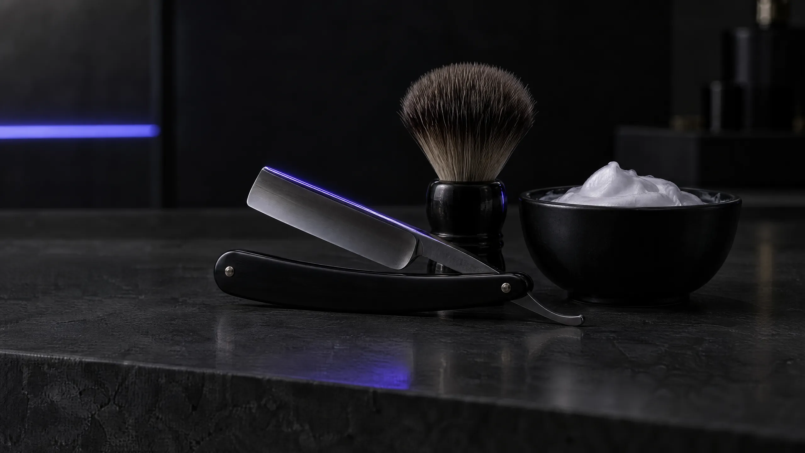 A straight razor, shaving brush and a bowl of lather on a dark counter